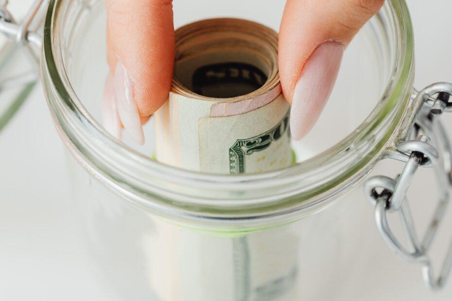 A careful hand storing a roll of banknotes inside a glass pot as a metafore of storing money safely